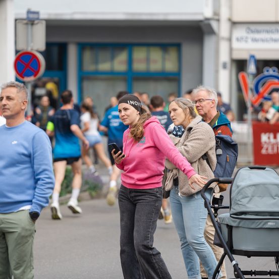 Eine Frau in einer pinkfarbenen Kapuzenjacke und ein Mann in einem blauen Shirt stehen neben einem Kinderwagen. Sie beobachten Läufer im Hintergrund.