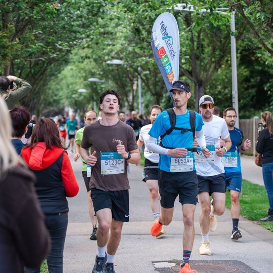 Marathonteilnehmer laufen auf einer von Bäumen gesäumten Straße. Ein Läufer mit blauem Shirt und der Nummer 5053A führt, gefolgt von anderen. Zuschauer säumen die Seiten, einige halten die Veranstaltung mit der Kamera fest.