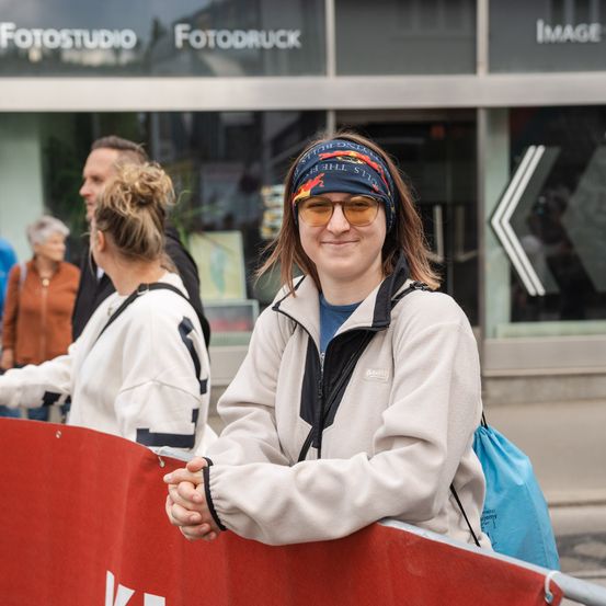 Eine Frau mit einer blauen Stirnband und einer Brille lächelt für ein Foto vor einem roten Barriere. Dahinter laufen Menschen an einem Gebäude mit dem Schild 'Fotostudio Fotodruck' vorbei.