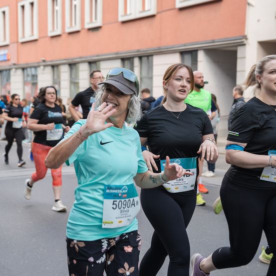 Eine Gruppe von Läufern in einem Marathon, zwei lächeln und gestikulieren, mit Gebäuden und anderen Läufern im Hintergrund.