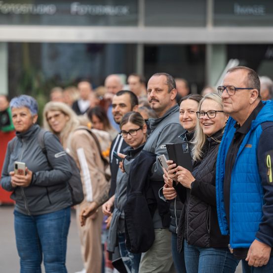 Eine Gruppe von Menschen steht in einer Reihe, einige halten Handys. Eine Frau mit Brille lächelt in die Kamera. Ein Mann in einer blauen Weste steht rechts.