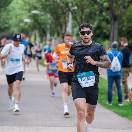 Eine Gruppe von Menschen nimmt an einem Marathon teil. Ein Mann vorne trägt die Nummer 7015C auf seinem Shirt. Unscharfer Hintergrund zeigt andere Läufer und Zuschauer.