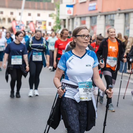 Eine Frau mit Brille geht mit Trekkingstöcken an einem Marathon teil. Sie trägt die Nummer 3001-02 und ein weiß-blaues Shirt. Hinter ihr laufen Menschen bei einem Marathon.