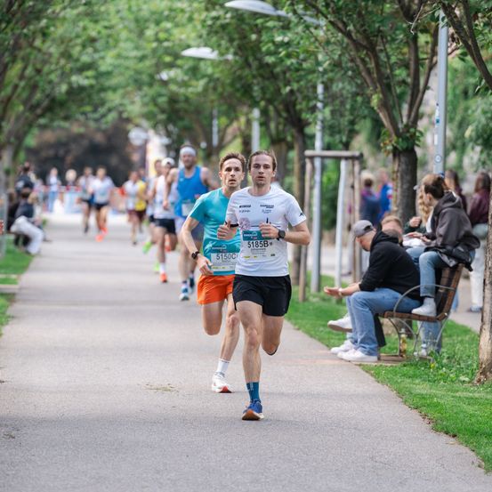 Mehrere Läufer nehmen an einem Marathon teil, einer führt in einem weißen T-Shirt, ein anderer in orangefarbenen Shorts. Zuschauer beobachten von den Seitenlinien, einige sitzen auf Bänken. Bäume säumen den Weg.