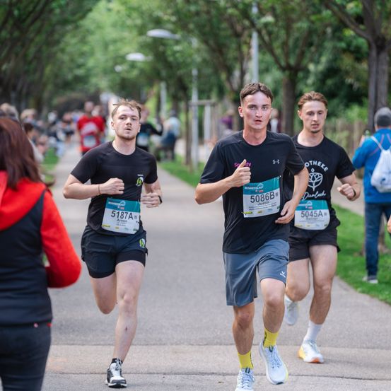Drei Männer laufen auf einem gepflasterten Weg, wahrscheinlich bei einem Marathon. Sie tragen schwarze T-Shirts mit Nummern. Bäume säumen die Seiten des Weges, und Menschen stehen auf dem Rasen.