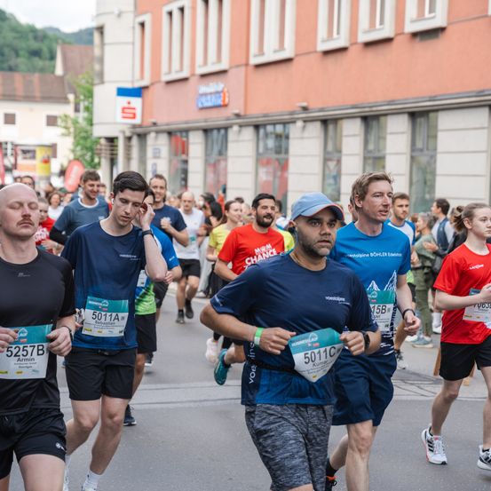 Läufer nehmen an einem Marathon auf einer Stadtstraße teil, mit Gebäuden und Zuschauern im Hintergrund. Mehrere Läufer tragen Startnummern und Namen.