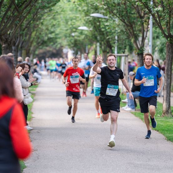 Läufer bei einem Marathon sprinten auf einem Weg, umgeben von Bäumen. Zuschauer feuern sie von den Seitenlinien an.