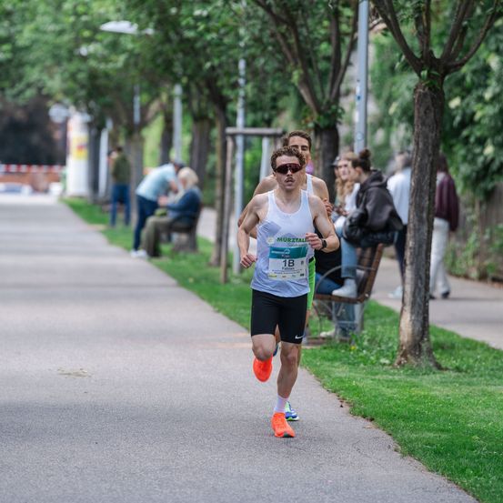 Ein Mann mit der Startnummer 1B läuft einen Marathon. Er trägt eine Sonnenbrille und Turnschuhe. Hinter ihm steht ein weiterer Mann mit der Hand auf seiner Schulter. Rechts sitzen Leute auf einer Bank.