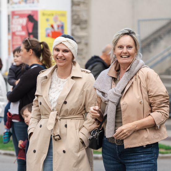 Zwei Frauen laufen lächelnd auf der Straße. Eine trägt einen beigefarbenen Trenchcoat und die andere einen beigefarbenen Mantel mit Schal. Hinter ihnen trägt eine Frau ein Baby und ein Mann steht neben einer Wand mit Postern.