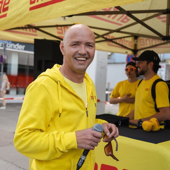 Ein Mann in einem gelben Hoodie lächelt und hält ein Mikrofon unter einem Zelt. Zwei Personen in gelben Shirts stehen hinter ihm. Ein schwarzer Tisch und ein Lautsprecher sind im Hintergrund.