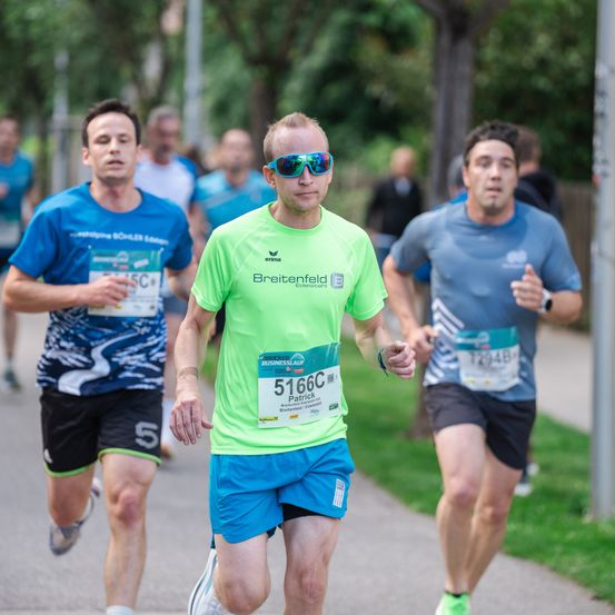 Ein Mann mit der Nummer 5166C auf seinem grünen Shirt läuft einen Marathon mit anderen Teilnehmern. Er trägt eine Sonnenbrille und blaue Shorts.