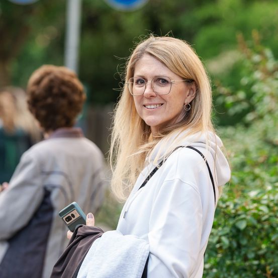 Eine Frau mit blonden Haaren, die eine Brille und eine weiße Kapuzenjacke trägt, hält ein Telefon und eine Jacke. Eine weitere Person in einer grauen Jacke ist im Hintergrund verschwommen.