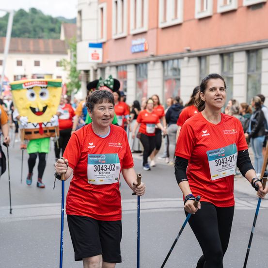 Zwei Frauen laufen mit Skistöcken bei einem Marathon. Eine trägt ein Shirt mit der Nummer 3043-02, und die andere hat 3043-01. Dahinter laufen Leute in Kostümen, darunter eine mit einem SpongeBob-Gesicht. Gebäude säumen die Straße.