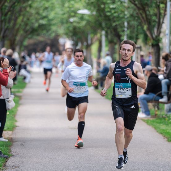 Zwei Marathonläufer laufen auf einem Betonweg, während Zuschauer sie beobachten. Der Läufer vorne trägt ein schwarzes Adidas-Shirt mit der Nummer 5270.