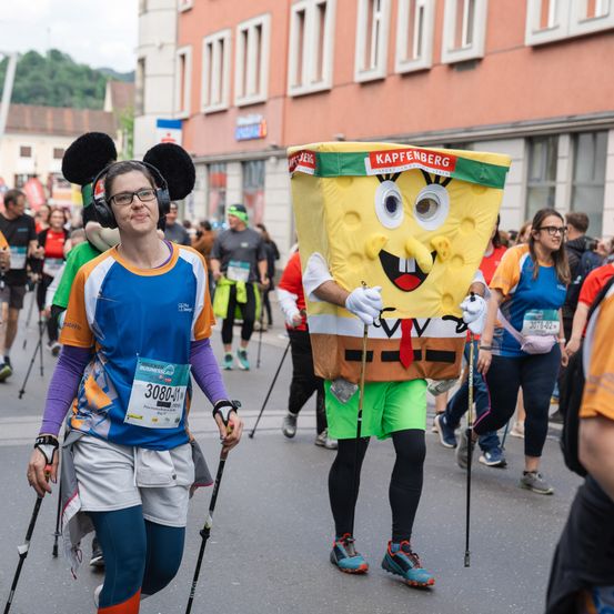 Ein Marathonläufer mit Micky-Maus-Kopfmaske und Kopfhörern läuft mit Trekkingstöcken. Ein Mann, verkleidet als SpongeBob Schwammkopf, folgt mit dem Startnummernband 308041. Sie laufen auf einer Straße mit anderen Läufern und Gebäuden.