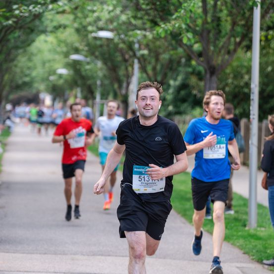 Eine Gruppe von Läufern nimmt an einem Marathon auf einer von Bäumen gesäumten Straße teil. Der Läufer im Vordergrund trägt ein schwarzes Shirt mit einem grünen Startnummer, gefolgt von einem Mann in einem roten Shirt.