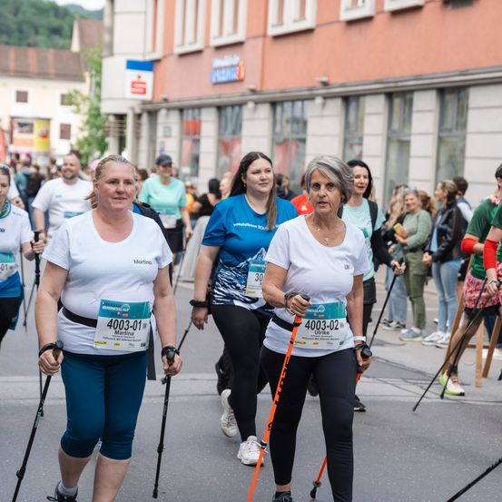 Eine Gruppe von Frauen nimmt an einem Rennen mit Gehstöcken teil. Sie tragen nummerierte Startnummern und T-Shirts. Die Frau in der Mitte hat ein blaues Shirt, die anderen tragen weiß. Im Hintergrund sind Gebäude zu sehen.