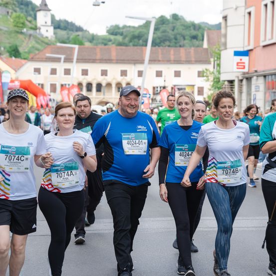Fünf Personen nehmen an einem Marathon teil, tragen nummerierte Trikots. Sie laufen in einer Reihe, mit einem großen Gebäude und Bergen im Hintergrund.