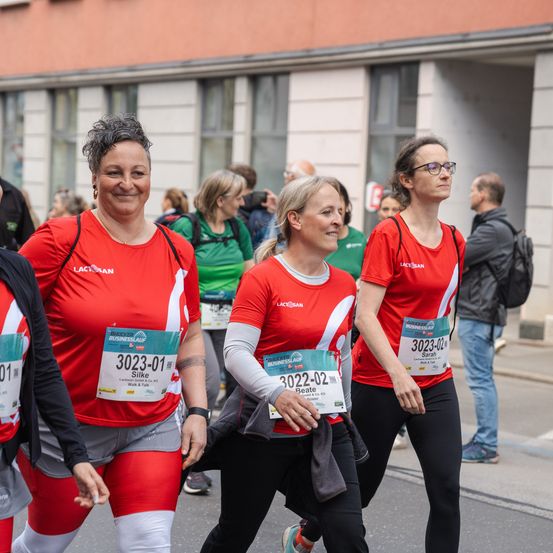 Drei Frauen in roter Laufausrüstung und Startnummern laufen bei einem Marathon mit. Ihnen folgt ein Mann mit Rucksack. Gebäude sind im Hintergrund.