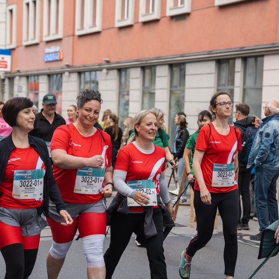 Vier Frauen in roten Laufoutfits nehmen an einem Marathon teil. Sie laufen auf einer Straße neben anderen Läufern. Dahinter beobachtet eine Menge von Menschen das Ereignis.