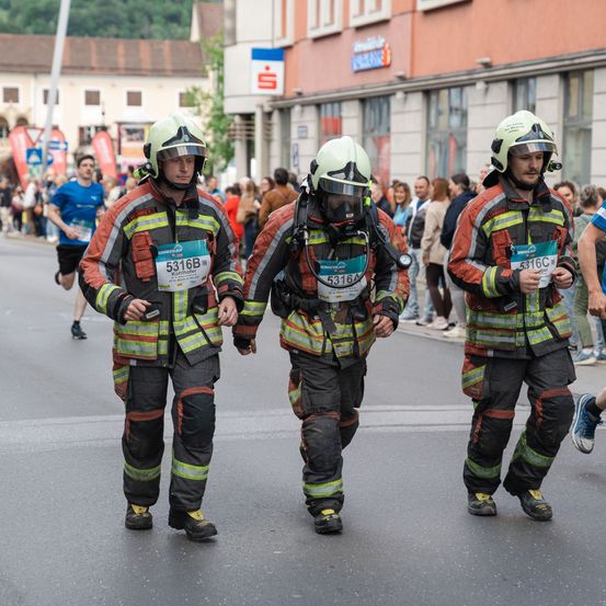 Drei Feuerwehrleute in Uniform laufen während eines Marathons auf einer Straße. Sie tragen Helme und Startnummern. Zuschauer stehen an den Seiten. Gebäude säumen die Straße.