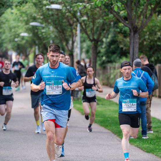 Eine Gruppe von Läufern bei einem Marathon, einer davon ist David mit der Nummer 7021B. Sie laufen auf einer Straße, gesäumt von Bäumen und Zuschauern.