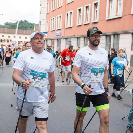 Zwei Männer nehmen an einem Nordic Walking Marathon teil. Beide tragen Startnummern, eine mit 3061-02 und die andere mit 3061-01. Sie gehen mit Stöcken auf einer Straße, auf der auch andere Teilnehmer unterwegs sind und Gebäude im Hintergrund stehen.