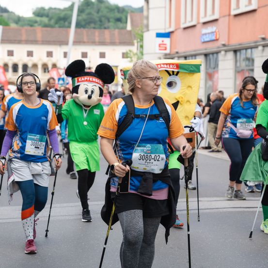 Eine Gruppe von Menschen nimmt an einem Marathon teil, darunter eine Frau mit Rucksack und ein Läufer in einer Micky-Maus-Kostüm. Zuschauer beobachten von den Seitenlinien.