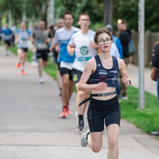 Eine junge Läuferin mit Brille nimmt an einem Marathon teil. Hinter ihr folgen mehrere andere Läufer. Im Hintergrund sind Bäume und ein Zaun zu sehen.