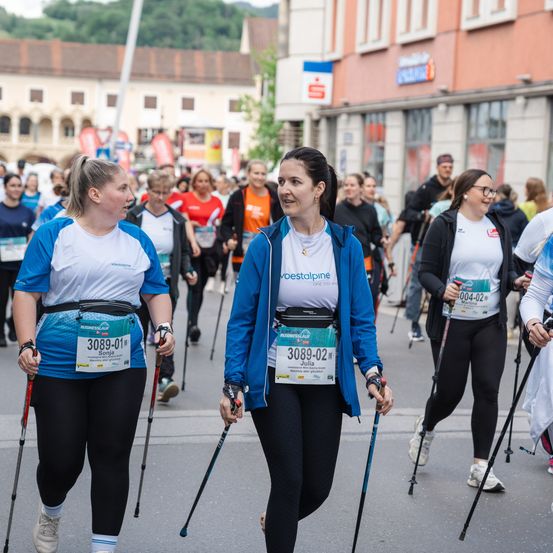 Mehrere Frauen mit Startnummern nehmen an einem Rennen teil. Sie benutzen Gehstöcke und sind von anderen Teilnehmern umgeben. Im Hintergrund sind Gebäude und Bäume zu sehen.