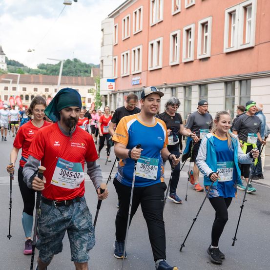 Eine Gruppe von Menschen in roten und blauen Shirts nimmt an einem Rennen teil. Sie tragen Skistöcke und laufen in einer Reihe auf einer Stadtstraße. Gebäude mit Fenstern säumen die Straße.
