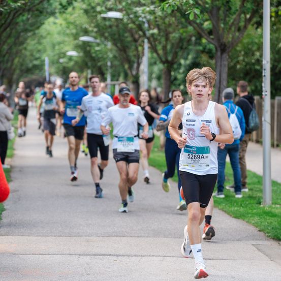 Ein Marathonrennen mit Läufern auf der Straße, einige tragen Startnummern und einer hat 5269A. Bäume und Straßenlaternen säumen den Weg.