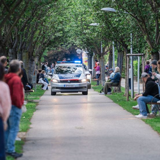 Ein Polizeiwagen fährt auf einem Bürgersteig mit Bäumen und Menschen auf beiden Seiten. Einige Leute sitzen auf Bänken und andere stehen.