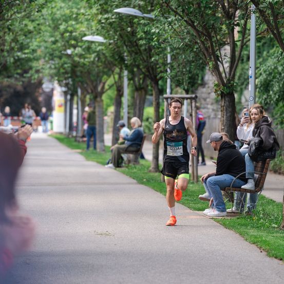 Ein Läufer bei einem Marathon mit der Nummer 5233 auf seinem Trikot sprintet auf einem von Bäumen gesäumten Weg, während Zuschauer zusehen und Fotos machen.