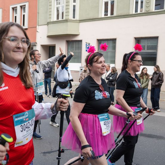 Eine Gruppe von Frauen läuft bei einem Marathon, zwei in Tüllröcken, und eine weitere Frau mit Brille beobachtet sie. Sie alle lächeln und halten Wanderstöcke. Im Hintergrund sind Gebäude und Menschen zu sehen.