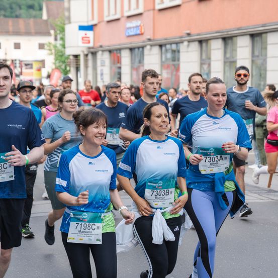 Eine Gruppe von Menschen läuft einen Marathon. Die Frau in der Mitte trägt die Nummer 7303B. Die Frau links von ihr trägt die Nummer 7198C. Dahinter trägt ein Mann die Nummer 7070. Gebäude befinden sich an der Seite.