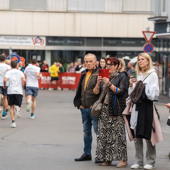 Eine Gruppe von Menschen steht auf einer Stadtstraße und beobachtet einen Laufwettbewerb. Ein Mann hält ein Handy und macht Fotos. Zwei Frauen, eine mit Brille, stehen neben ihm und machen auch Fotos.