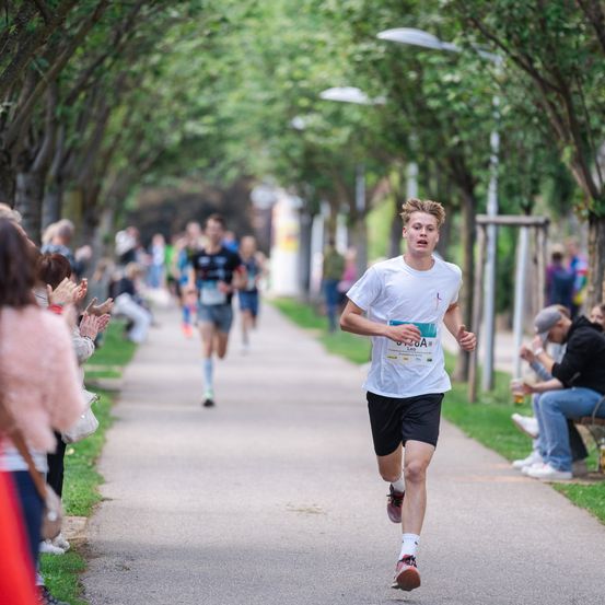 Ein junger Läufer nimmt an einem Marathon bei sonnigem Wetter teil, während Zuschauer an der Seite ihn anfeuern.