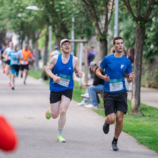 Zwei Männer laufen bei einem Marathon auf einer von Bäumen gesäumten Straße. Einer hat eine weiße Kappe und ein blaues Tanktop, der andere trägt ein blaues T-Shirt. Zuschauer beobachten sie.