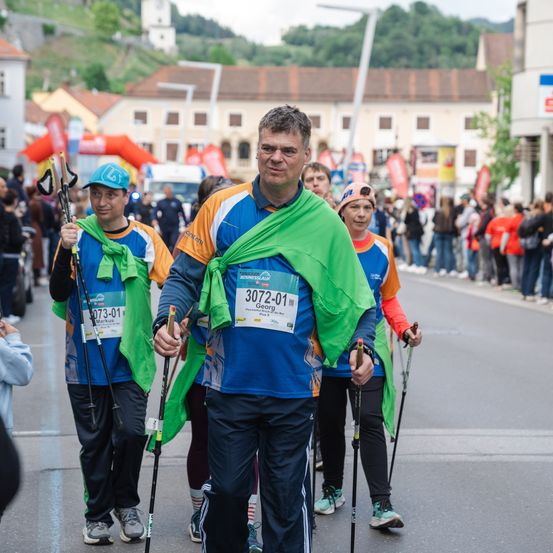 Läufer nehmen an einem Marathon teil, wobei ein Mann vorne ein Startnummer 3072-01 trägt. Ihnen folgen eine Menge und Gebäude.