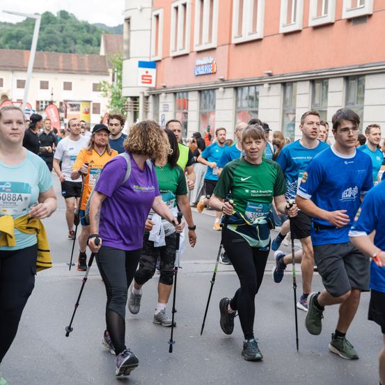 Eine Gruppe von Marathonläufern, darunter eine Frau mit einer Prothese, nehmen an einem Rennen teil. Sie laufen auf einer Stadtstraße, mit Gebäuden im Hintergrund.