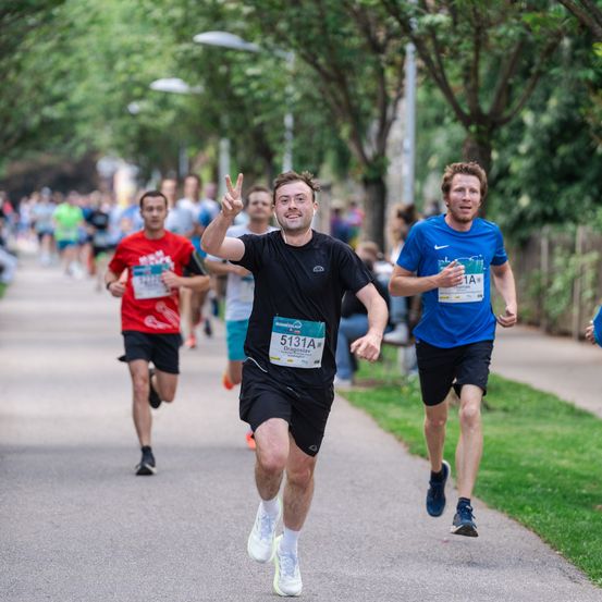 Läufer nehmen an einem Marathon auf einer asphaltierten Straße mit Bäumen und Grünflächen teil. Ein Mann in der Mitte winkt, während andere hinter ihm laufen.