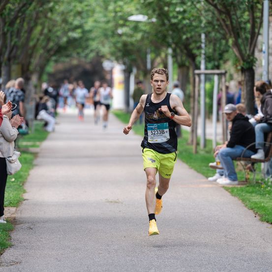 Ein männlicher Läufer in einem schwarzen Tanktop und gelben Shorts läuft einen Marathon. Zuschauer beobachten ihn am Straßenrand.