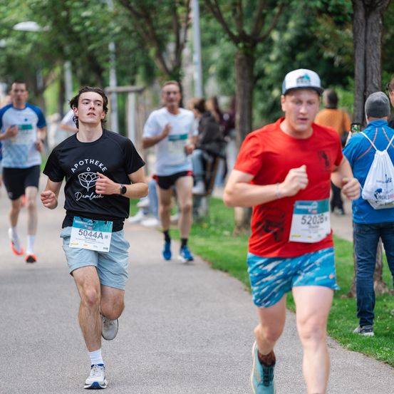 Läufer nehmen an einem Marathon teil. Ein Mann in einem schwarzen T-Shirt mit der Nummer 5044A läuft. Ein anderer Läufer trägt ein rotes Shirt und blaue Shorts. Zuschauer sind an der Seite.