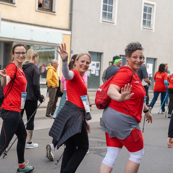 Drei Frauen in roter Laufausrüstung nehmen an einem Marathon teil, winken und lächeln. Andere Läufer, Zuschauer und ein Gebäude mit einem gerahmten Fenster sind im Hintergrund zu sehen.