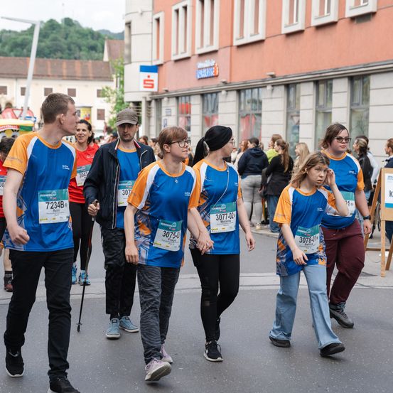 Eine Gruppe von Marathonläufern, die blaue und weiße Shirts tragen, geht auf der Straße. Hinter ihnen befindet sich ein Gebäude mit einem Schild mit einem Logo.