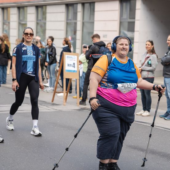 Eine Frau mit einem Lächeln auf dem Gesicht läuft in einem Marathon mit der Startnummer 2079-01. Sie trägt Kopfhörer und hält einen Gehstock. Dahinter läuft eine Frau auf der Straße.