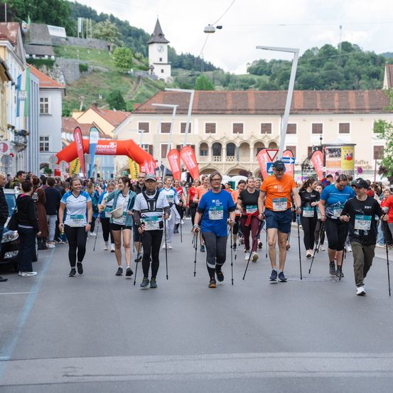Eine Gruppe von Menschen mit Trekkingstöcken läuft in einem Marathon. Sie tragen nummerierte Startnummern und werden von Zuschauern umgeben. Gebäude säumen die Straße.