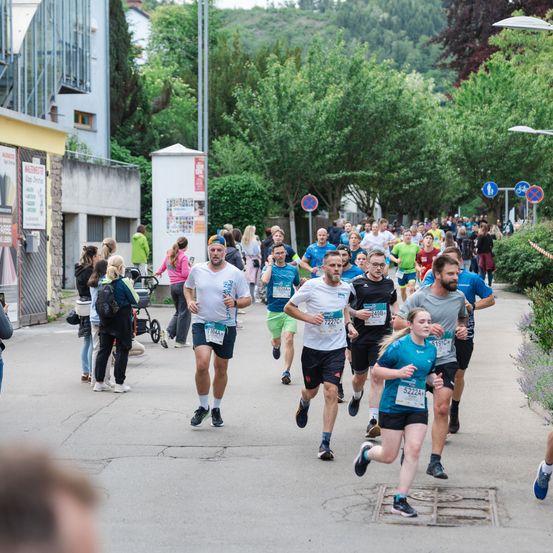 Eine Gruppe von Menschen läuft in einem Marathon auf einer Stadtstraße mit Bäumen, einem Gebäude und Straßenschildern. Zuschauer stehen in der Nähe.