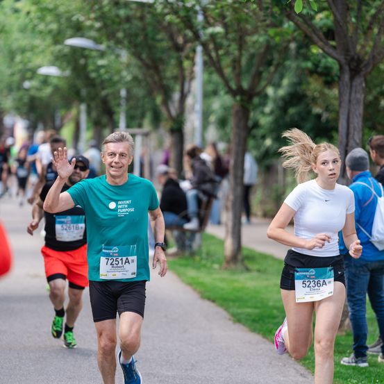 Ein Mann und eine Frau laufen in einem Marathon. Der Mann winkt, während die Frau vorausläuft. Zuschauer säumen die Straßenseiten.